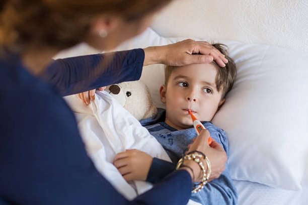 Sick boy with thermometer laying in bed and mother hand taking temperature. Mother checking temperature of her sick son who has thermometer in his mouth. Sick child with fever and illness while resting in bed.