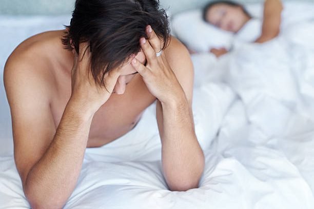 A young man sitting on the edge of his bed while his wife sleeps in the background